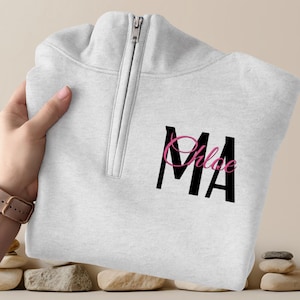 Puede incluir: Una sudadera gris claro con cremallera de cuarto de cremallera con las letras "MA" en negro y el nombre "Chloe" en escritura rosa. La sudadera tiene una cremallera plateada y está doblada. El fondo es beige neutro.