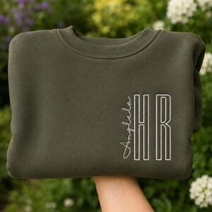 Peut inclure: Un sweat-shirt à col rond vert olive avec une broderie blanche sur le côté droit. Le motif comprend le prénom Angela écrit en écriture cursive verticalement, à côté des grandes lettres capitales HR en gras et contournées.