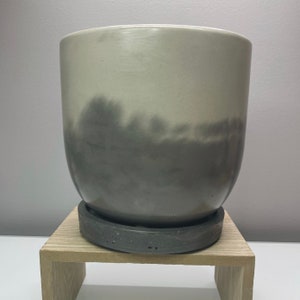 May include: A gray and white ceramic planter with a subtle ombre design. The planter has a round shape and a wide base.