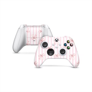 May include: Two white Xbox controllers with a pink and white striped design. The design includes pink bows. The controllers are set against a white background.