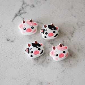 May include: Four white cow-shaped charms with pink and black markings. Each charm has a pink nose, black eyes, and small horns. The charms are arranged on a light gray surface.