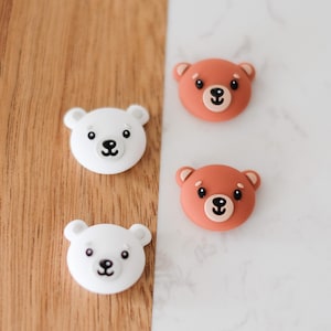May include: Three silicone bear face charms, two white and one brown. The white bears have black eyes and noses, and the brown bear has pink eyes and nose.