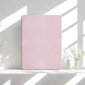 Puede incluir: Un objeto rectangular rosa claro con esquinas redondeadas, posiblemente una almohadilla o una cubierta, está centrado en la imagen. El objeto tiene una superficie texturizada. El fondo es una pared blanca con sombras y pequeños jarrones con flores blancas.