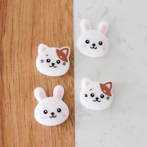 May include: Four silicone charms featuring cartoon animal faces. Two charms are white bunnies with pink ears and black eyes. Two charms are white cats with brown ears and a brown patch on their head.