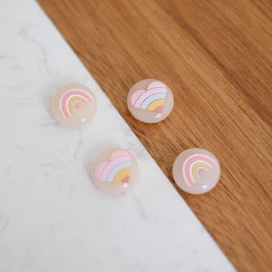 Puede incluir: Cuatro imanes decorativos redondos, de color rosa pastel, con diseños de arcoíris y corazón.