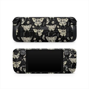 May include: A black handheld gaming console with a cream-colored moth and floral pattern. The console has a black screen and black buttons. The design includes moths, flowers, and stars. The word "STEAM" is visible on the bottom left.