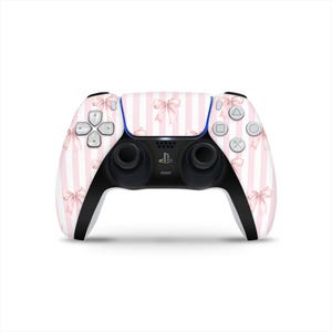Puede incluir: Un mando de PlayStation 5 a rayas blancas y rosas con un patrón de lazos. El mando tiene asas y botones negros con un acento azul. El logotipo de PlayStation está centrado en la parte inferior.