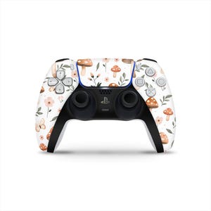 Könnte beinhalten: Ein weißer PlayStation 5-Controller mit einem Blumen- und Pilzmuster. Das Design umfasst braune Pilze, rosa Blüten und grüne Blätter. Der Controller hat schwarze Griffe und Tasten mit einem blauen Akzent.