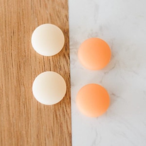 May include: Four round, smooth objects. Two are a creamy off-white colour, and two are a vibrant orange. The objects are arranged on a wooden surface and a white marble surface.