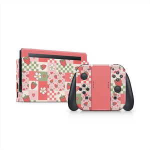 Puede incluir: Una consola y mandos Nintendo Switch rosas con un estampado de fresas y flores. El diseño presenta fresas, flores y corazones sobre un fondo de patchwork de cuadros rosas, verdes y blancos. Los mandos tienen detalles en negro.