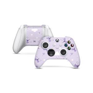 Puede incluir: Un mando de Xbox morado con un patrón de mariposas y flores. El mando tiene un fondo blanco con mariposas moradas y flores blancas.