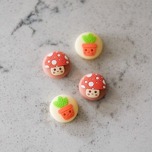 May include: Four round, decorative buttons with cartoon mushroom and potted plant designs. The mushroom buttons have red caps with white spots and smiling faces. The plant buttons feature a smiling orange pot with green leaves.