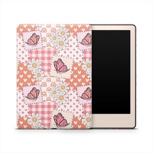 May include: A decorative e-reader cover featuring a patchwork design with pink butterflies, white daisies, rainbows, and hearts on a peach background. The cover is paired with a beige e-reader device.