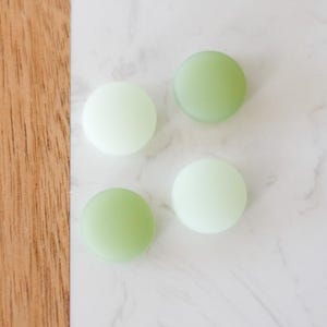 May include: Four round, light green and white objects are arranged on a white marble surface with a wooden edge. The objects have a smooth, matte finish and are evenly spaced, creating a simple, minimalist aesthetic.