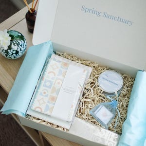May include: Open gift box with the text "Spring Sanctuary" on the lid. Inside, a patterned notebook, a candle, and small square items in a blue organza bag rest on shredded paper. A decorative glass orb and diffuser are also visible.