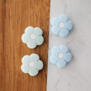 以下が含まれることがあります： 花の形をしたシリコン製の引き出しノブが4つ、薄い青が2つ、薄い青が2つ。ノブの中央には白い円があります。