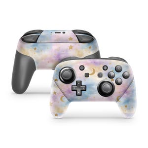 May include: Two wireless video game controllers featuring a pastel watercolor pattern in shades of blue, purple, and yellow. The surface is decorated with small gold stars and crescent moons. The controllers have black buttons, joysticks, and triggers.