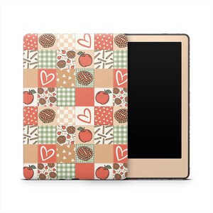 May include: A tan e-reader with a patterned cover. The cover features a patchwork design with red apples, pie slices, hearts, and green gingham squares. The e-reader has a black screen and a gold-colored frame.