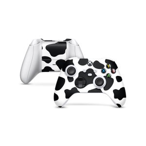Può includere: Due controller Xbox bianchi con un design a stampa di mucca in bianco e nero. I controller hanno pulsanti neri e pad direzionali. Il logo Xbox è visibile sui controller.