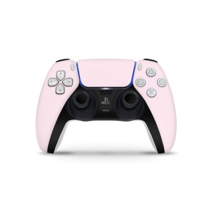 Puede incluir: Un mando inalámbrico PlayStation 5 DualSense rosa con detalles negros y botones blancos. El mando tiene un logotipo de PlayStation en la parte delantera.