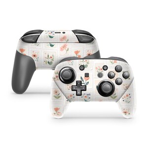 May include: Two wireless video game controllers featuring a light-colored skin with a repeating pattern of small, rectangular postage stamp-style illustrations. Each stamp displays various delicate floral motifs in shades of peach, blue, and green.