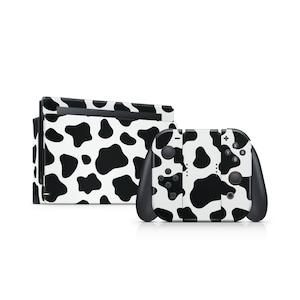May include: A white and black cow print skin set for a gaming console. The set includes a console and two controllers. The controllers are black with white and black cow print designs on the front. The console is white with black cow print.