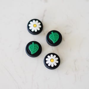 May include: Four round black silicone controller caps with different designs. Two caps feature a white daisy with a yellow centre. Two caps feature a green leaf.