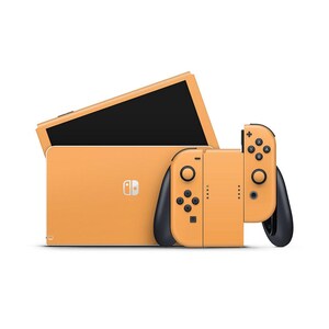 May include: An orange video game console with black accents. The console includes a screen, a main unit, and two detachable controllers. The controllers have black joysticks and buttons. The console is set against a white background.