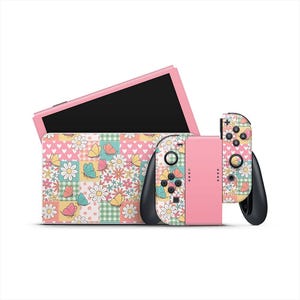 Puede incluir: Un conjunto de consola y controlador Nintendo Switch rosa. El controlador y la funda presentan un diseño de patchwork con mariposas, margaritas y corazones. La consola es rosa con una pantalla negra.