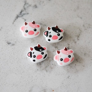 May include: Four small silicone cow charms in white, black, and pink. The cow charms have black eyes, pink noses, and brown horns.