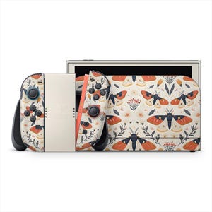 Peut inclure: Une console Nintendo Switch et ses manettes, sur fond crème. Les manettes et la console présentent un motif répété de papillons de nuit, de fleurs et de feuilles en orange, noir et crème. La Switch a des boutons noirs et un écran crème.