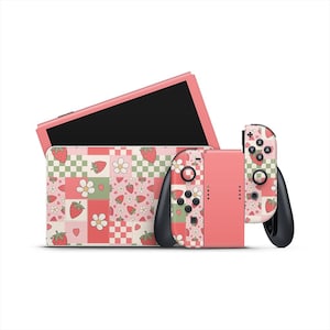 Puede incluir: Una consola Nintendo Switch rosa con una funda y mandos a juego. La funda y los mandos presentan un diseño de patchwork con fresas, flores y corazones. La pantalla es negra.