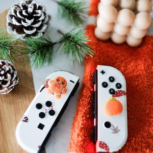 以下が含まれることがあります： お祝いの装飾が施された2つの白いNintendo Switch Joy-Con。片方にはジンジャーブレッドマン、もう片方にはカボチャが描かれています。どちらも黒いボタンがあり、秋と冬をテーマにしたグラフィックで装飾されています。コントローラーは、松の葉と質感のあるオレンジ色の表面を背景に配置されています。