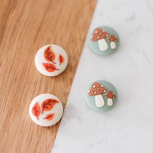 May include: Four decorative round buttons. Two buttons feature orange and red autumn leaves on a white background. The other two buttons have a light green background with a mushroom design in brown and white.