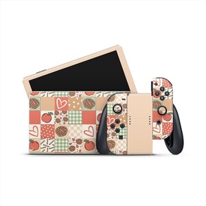 Puede incluir: Una consola Nintendo Switch beige con una funda y mandos estampados. La funda y los mandos presentan un diseño de patchwork con gráficos de manzanas, corazones y tartas en rojo, verde y beige. La pantalla de la consola es negra.
