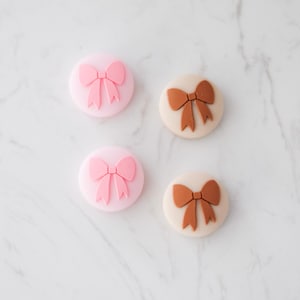 Op de afbeelding: Vier ronde siliconen knoppen met een reliëf ontwerp van een strik. Twee knoppen zijn roze met een roze strik en twee knoppen zijn beige met een bruine strik.