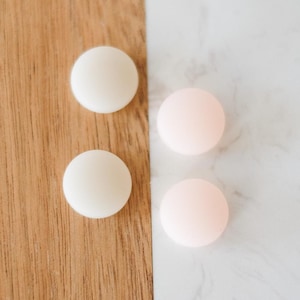 Puede incluir: Vista superior de cuatro objetos redondos y lisos. Dos son de color blanco crema y dos de color rosa pálido. Están dispuestos sobre un fondo dividido de madera y mármol blanco.