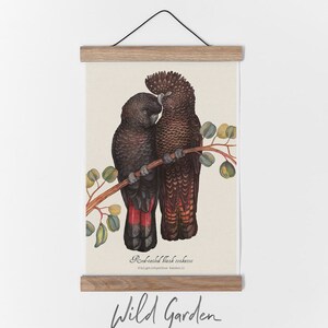 May include: A framed print featuring two Red-tailed Black Cockatoos perched on a branch with green leaves. The birds have black and red plumage. The print is held by wooden bars and a black string. The words "Wild Garden" are written at the bottom.