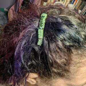 Puede incluir: Una pinza para el pelo verde y negra con un patrón moteado. La pinza está hecha de un material natural y tiene una forma única y orgánica.