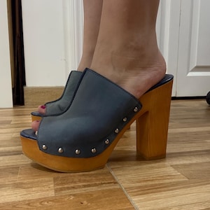 May include: Blue leather platform clogs with wooden heels. The open-toe mules feature a blue upper with silver stud accents and a light brown wooden platform and heel. The clogs have a high heel.