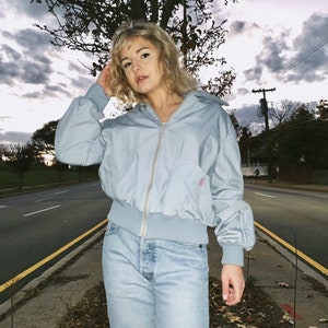 May include: A light blue bomber jacket with a zipper closure and ribbed cuffs and hem. The jacket is paired with light wash denim jeans. The background features a road, trees, and a cloudy sky.