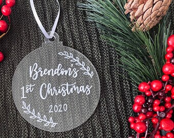 Puede incluir: Adorno de Navidad de acrílico transparente con letras blancas que dicen "Brendon's 1st Christmas 2020" con un diseño de hojas alrededor del texto.