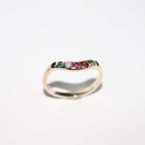 Puede incluir: Un anillo de banda de oro con un diseño ondulado y una fila de piedras preciosas de colores. Las piedras preciosas son rojas, verdes, moradas, blancas y azules.