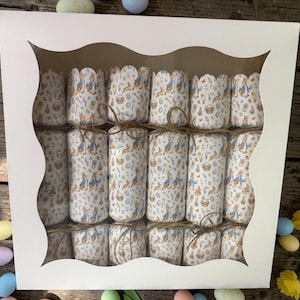 May include: A white box with a scalloped window displays twelve Easter crackers. Each cracker is cylindrical, wrapped in paper with a Peter Rabbit design, and tied with twine. The crackers are arranged in three rows of four.