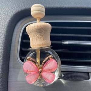 May include: A car air freshener with a clear glass bottle and a pink butterfly design. The butterfly has pink translucent wings and gold-colored accents. The top is made of light-coloured wood.