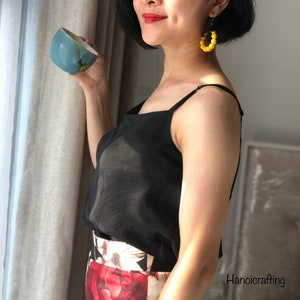 Pode incluir: Uma mulher vestindo um top preto sem mangas e uma saia floral com um brinco de argola amarelo com contas. Ela está segurando uma xícara de cerâmica azul e branca.
