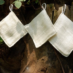 May include: Three square, off-white fabric items with hanging loops are displayed against a tree trunk. The items have a textured appearance and are likely handmade. The neutral color and simple design suggest a focus on natural materials.