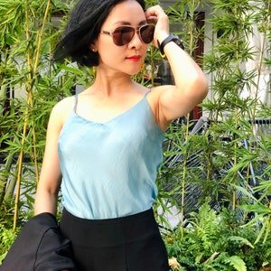 May include: A woman wearing a light blue tank top and black pants stands in front of a green leafy background. She is wearing sunglasses and has her hand on her head.