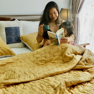 May include: A woman and a child are sitting on a bed with a yellow quilted duvet. The woman is holding a cup and a book, while the child is looking at the book. The bed has white pillows with a blue patterned pillow in the middle.