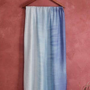 Könnte beinhalten: Ein blau-weißer Ombre-Schal hängt an einem Holzbügel vor einem roten Hintergrund. Der Schal geht von Weiß über Hellblau zu Dunkelblau über. Das Handy Silk-Logo ist unten rechts sichtbar.